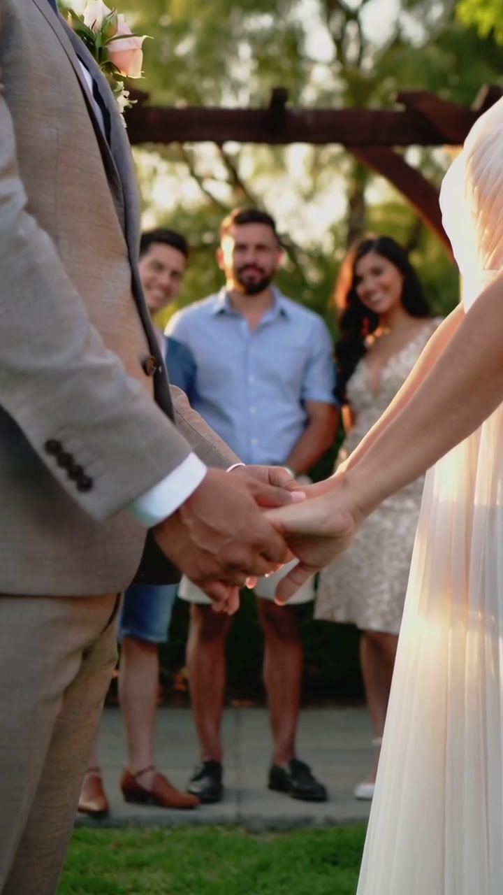 Vertical video: Holding bride and groom saying vows in yard, clasped hands, boutonniere, pale dress