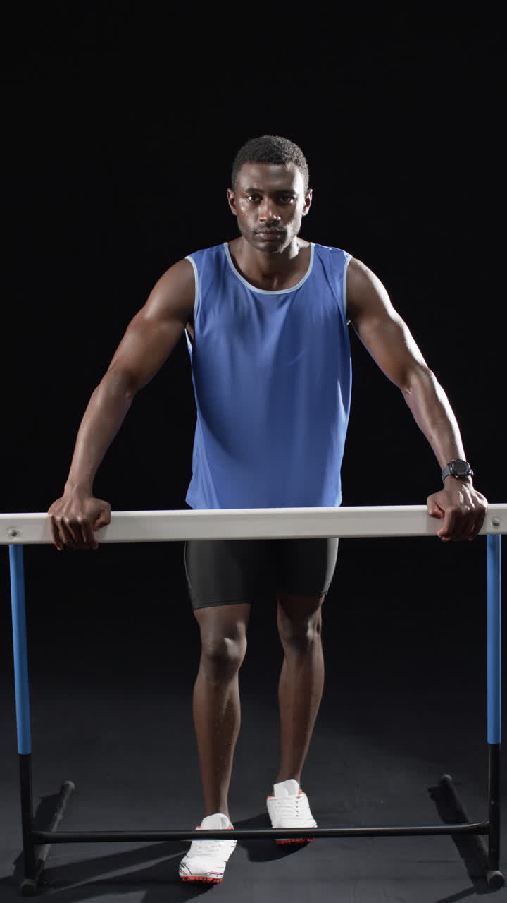 Vertical video: African American male athlete holding onto horizontal hurdle, black background