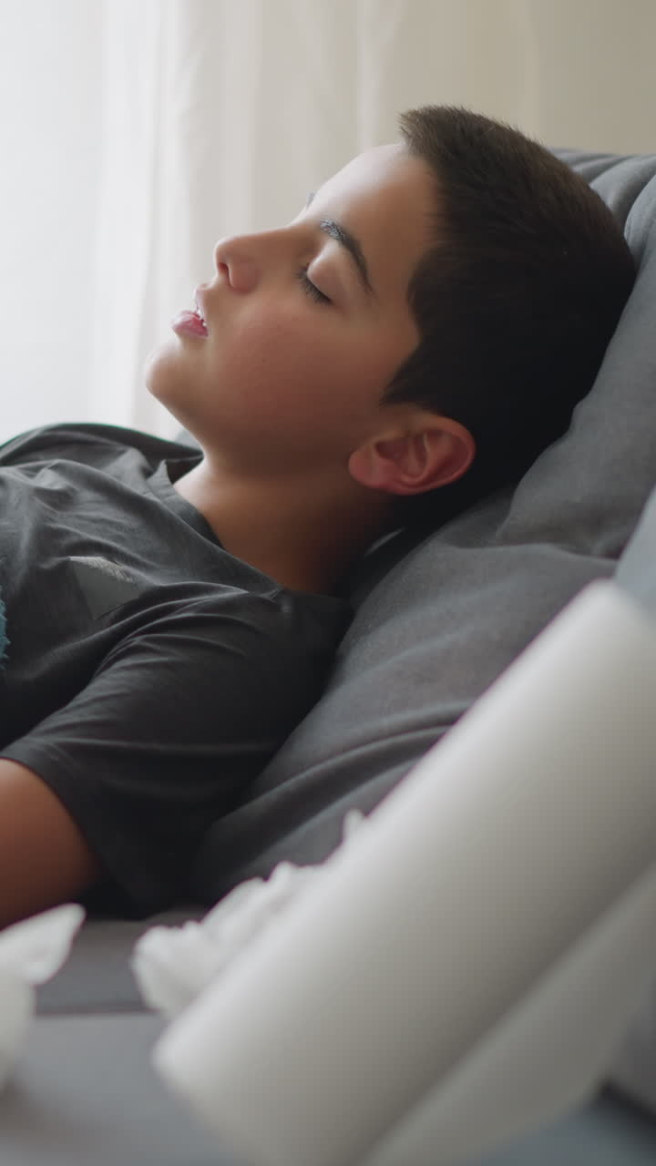Side view of boy resting on couch wrapped in blanket adjusting for comfort with used and unused tissue by his side, peaceful indoor setting with soft natural light coming through window