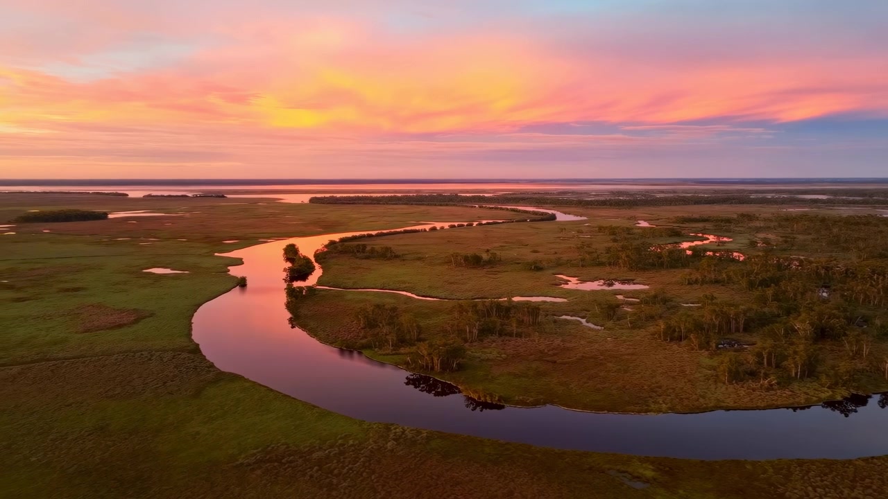 Un fiume serpeggiante si snoda attraverso una pianura paludosa erbosa punteggiata da piccole isole di alberi, che riflette un cielo al tramonto rosa-arancio.