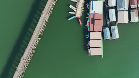 The-iconic-Mon-Bridge-and-a-harbor-with-colorful-boats-on-the-Khwae-Noi-River-in-Songklaburi,-Thailand,-embodying-the-vibrant-charm-of-the-region