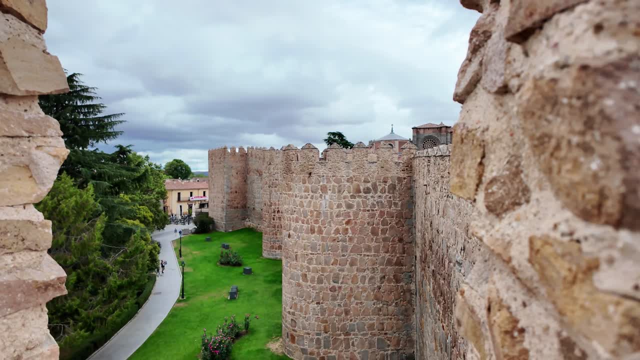 Vidvinkelbild av de historiska medeltida stadsmurarna i Ávila, Spanien, som sträcker sig över en gräsbevuxen kulle under en dramatisk molnig himmel. En av Europas bäst bevarade befästa städer och en UNESCO-världsarvsplats.