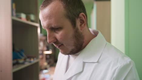 a close-up view of a man wearing a white lab coat walking inside his lab, with a shelf behind him with working tools in the shelf and wall painted green