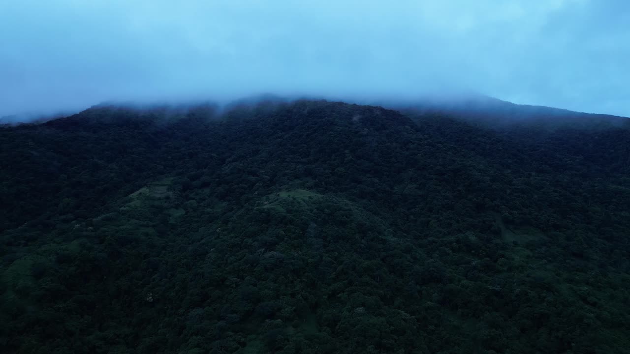 A pitch drone shot tilting down over a mist-covered tropical mountain, revealing dense green forest and rolling hills under a moody overcast sky of Mount Banahaw, Quezon Province Philippines