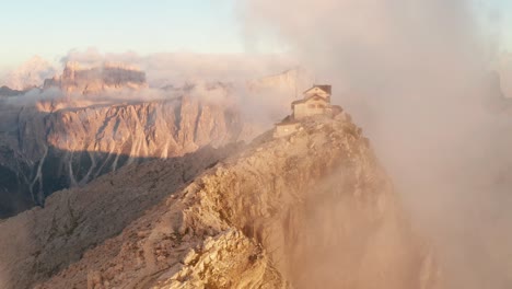 Mysterious-clouds-envelop-Nuvolau-mountain-lodge-illuminated-by-sunset---aerial-pullback-reveals-scenic-setting-atop-mountain-peak-in-Dolomites