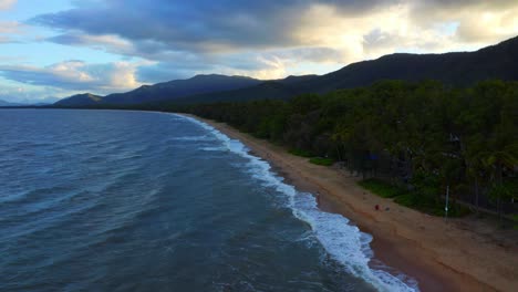 Vista-Aérea-De-Las-Olas-Del-Mar-De-Coral-Salpicando-En-La-Playa-De-Arena-En-Palm-Cove,-Región-De-Cairns,-Queensland,-Australia-Con-Cielo-Nublado