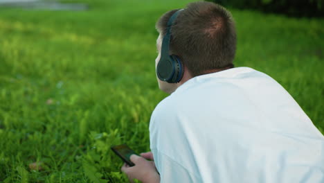 back view of man lying on grassy field wearing headphones, holding smartphone, deeply immersed in his device nods his head to the music, surrounded by lush green grass