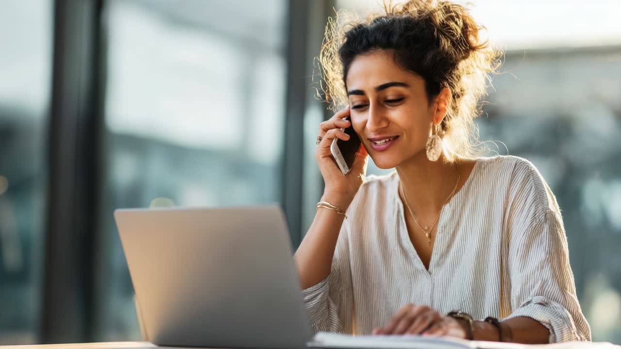Una giovane donna impegnata in una conversazione produttiva sorride mentre usa il suo portatile e parla al telefono, illuminata dalla calda luce naturale in un ambiente di lavoro moderno