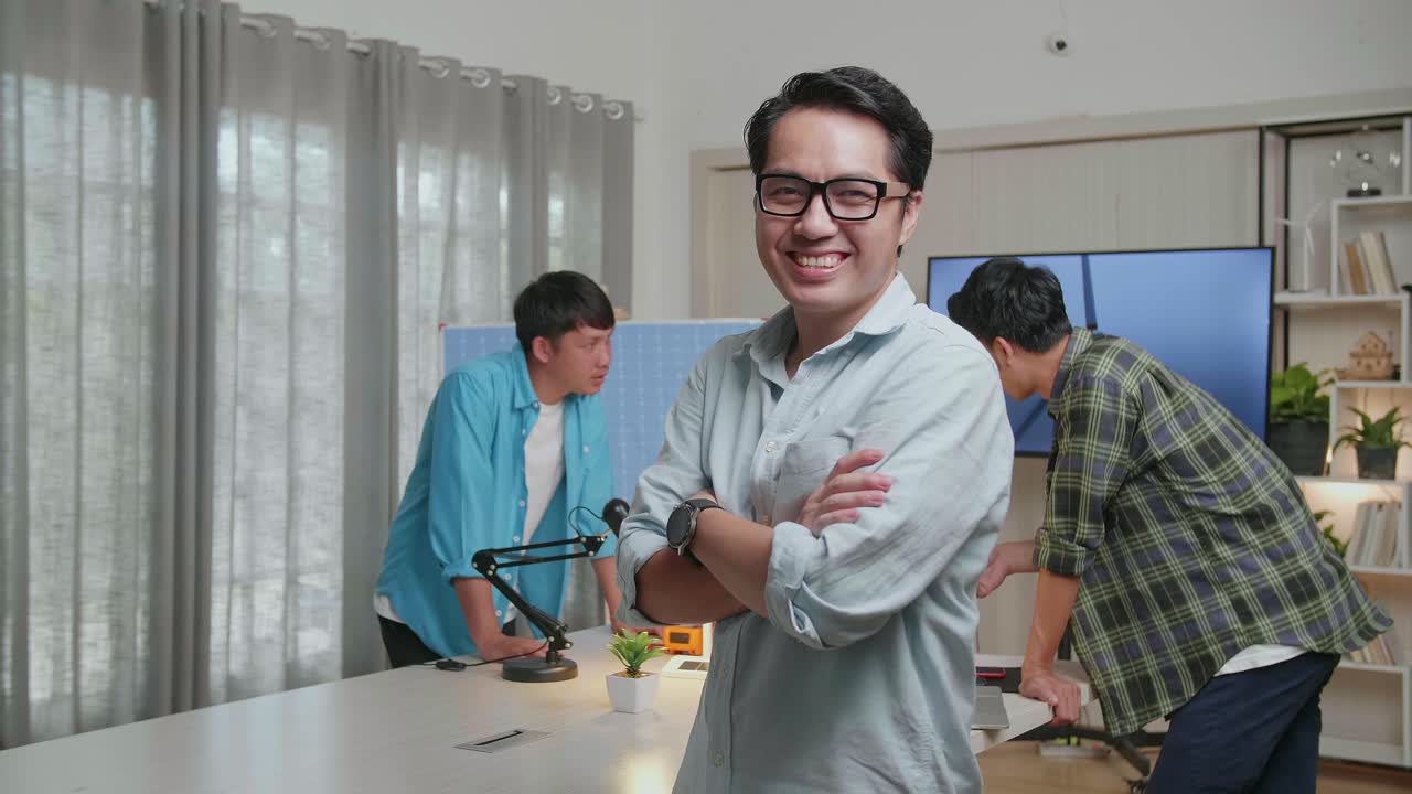 동료들이 사무실에서 풍력 터빈에 대해 논의하는 동안 미소 짓는 아시아 남자 엔지니어가 카메라에 팔을 고 있습니다.