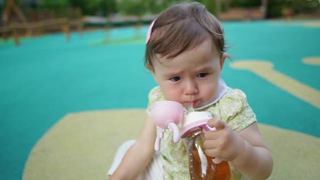 Porträt-Eines-1-jährigen-Süßen-Babys,-Das-Saft-Aus-Der-Flasche-Trinkt-Und-Ihn-Mit-Einer-Hand-Hält,-Wenn-Er-Auf-Einem-Spielplatz-Im-Freien-Sitzt