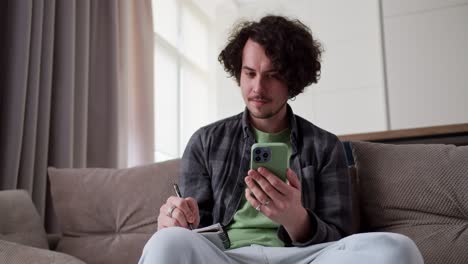 Confident-brunette-guy-with-a-mustache-in-a-checkered-gray-shirt-watches-a-video-on-a-green-smartphone-and-records-important-moments-while-sitting-on-the-sofa-at-home