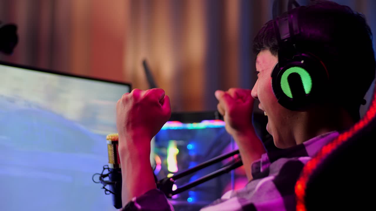 detailní pohled zezadu na asijského streamera, jak křičí gól a tančí, slaví vítězství v automobilové závodní hře na osobním počítači. Živý přenos videohry, stůl osvětlený RGB LED páskem
