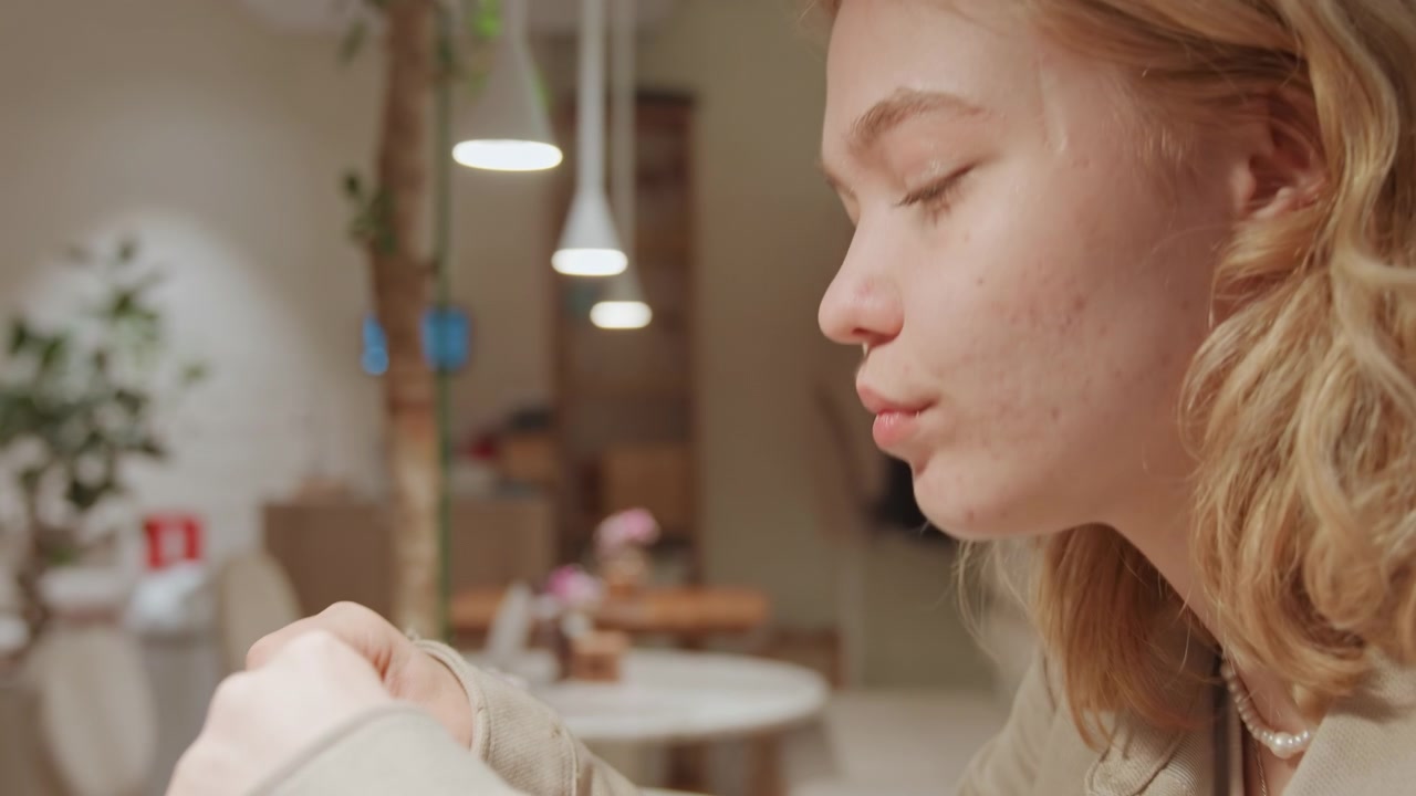 Primo piano di una giovane donna che mangia a un tavolo di un caffè, con illuminazione soffusa e piante da interno. La donna tiene uno snack tra le mani, indossando abiti casual, mentre è seduta all'interno. Lo sfondo presenta.
