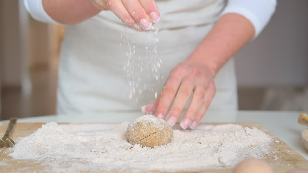 Seorang wanita mengenakan celemek, menambahkan tepung ke adonan di atas talenan kayu di dapur