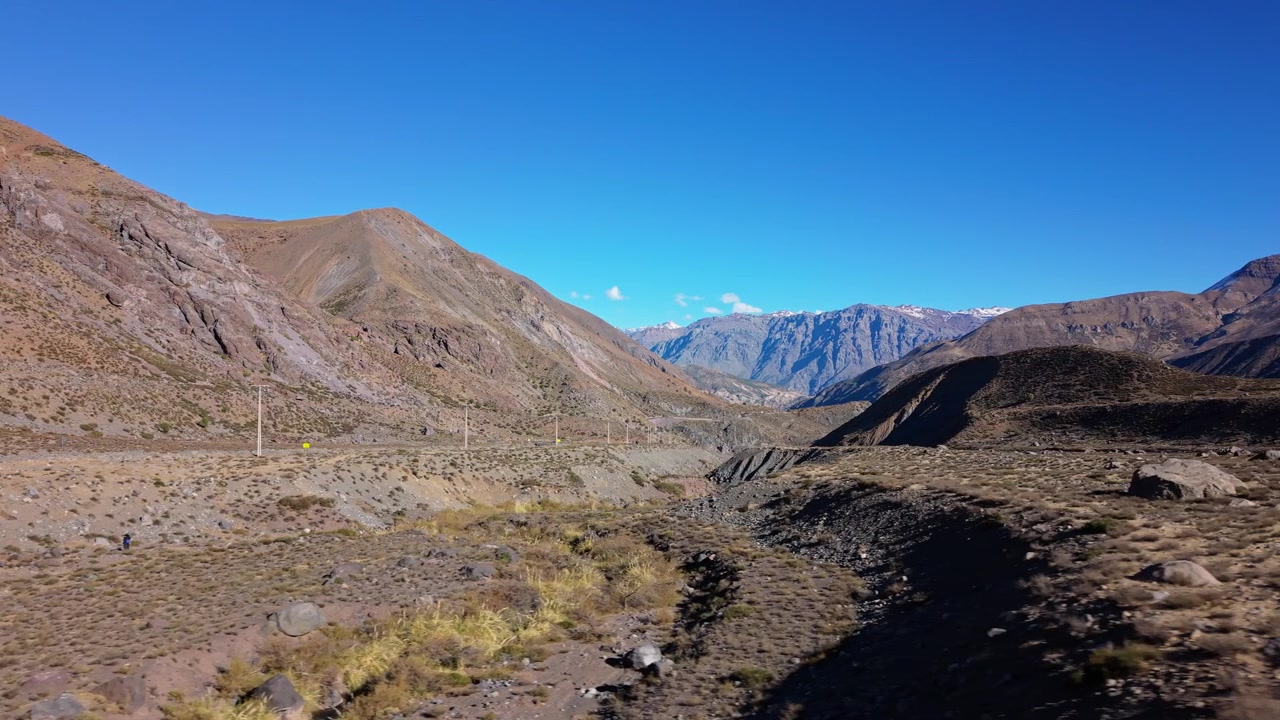 Movimento laterale basso lungo una strada polverosa di una valle di montagna, delimitata da pendii rocciosi aridi e vegetazione sparsa, con lontane cime andine innevate che si ergono all'orizzonte lontano e limpido