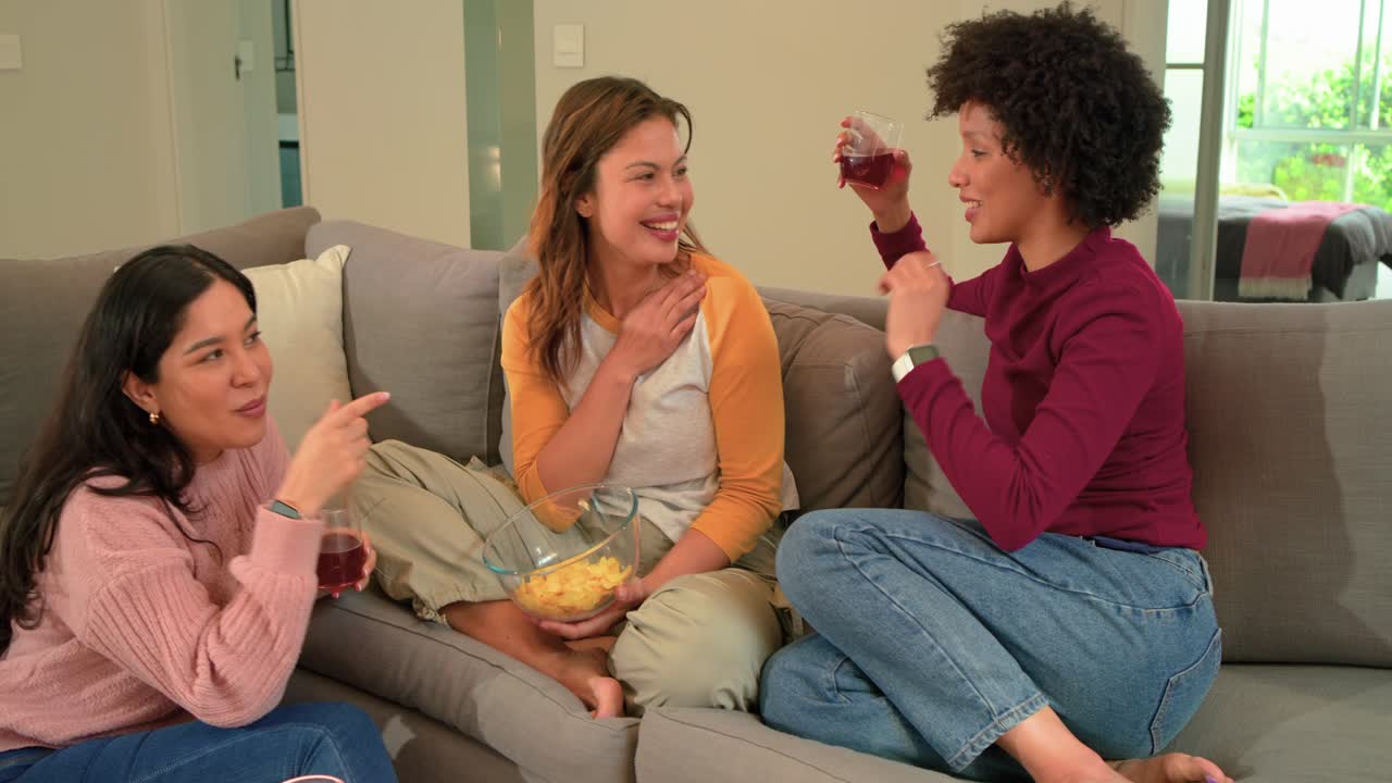 Amiche diverse che reagiscono a una battuta, fanno tintinnare i bicchieri, condividono patatine e ridono sul divano
