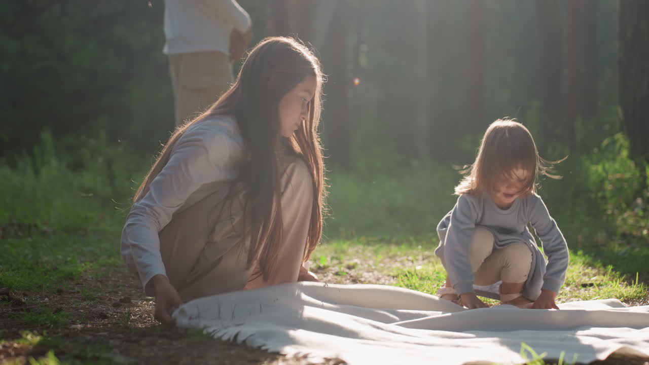 Young girl adjusting blanket carefully on green forest ground as sister approaches to help, sunlight illuminating scene with soft warmth, showing family bonding, and peaceful outdoor preparation