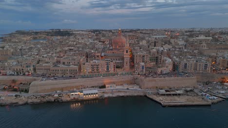 Una-Vista-Aérea-De-La-Antigua-Ciudad-De-La-Valeta-En-Malta-Al-Anochecer,-Cuando-Comienza-A-Iluminarse-Por-La-Noche,-Mostrando-La-Basílica-De-Nuestra-Señora-Del-Monte-Carmelo.