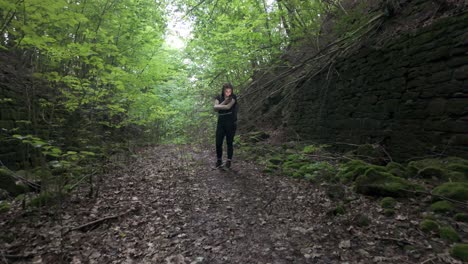 Mujer-Joven-Parada-En-El-Bosque,-Revisando-Mensajes-En-Su-Teléfono,-En-Medio-De-La-Saturación-Tecnológica-Incluso-En-La-Naturaleza,-Destacando-La-Dependencia