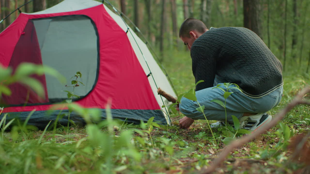 man squats next to red tent using thick stick to drive peg into grassy forest floor surrounded by green plants and trees focused on camping task during quiet outdoor moment in wilderness