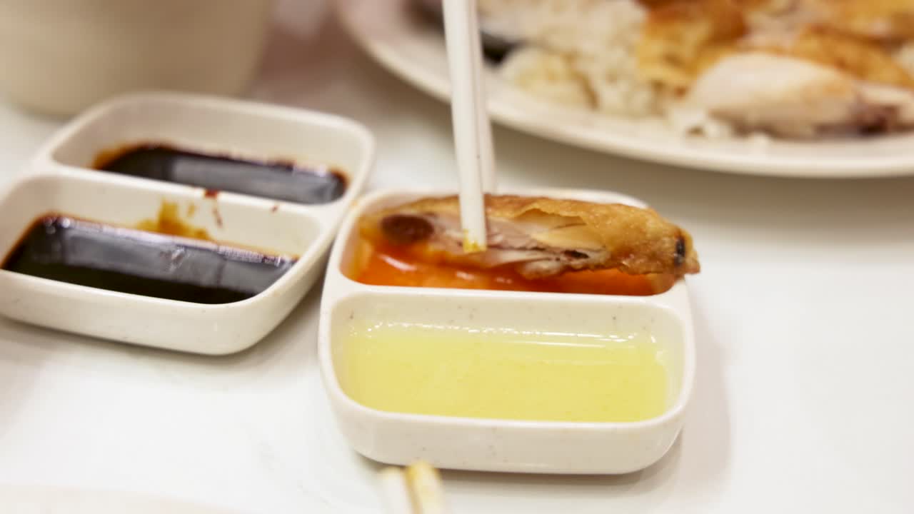 Chopsticks pick up roasted chicken and dip it into vibrant sauces beside Hainanese chicken rice, under bright indoor lighting with a steady close-up camera angle