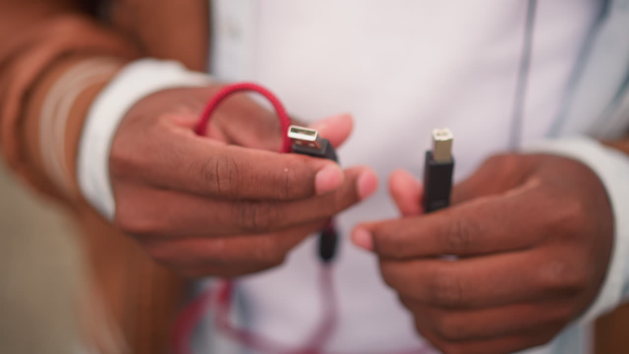 black busker hands plugging red cable for street performance, intimate closeup of USB and jack on braided cord, casual jacket and denim, testing gear and tuning before busking set
