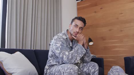 thoughtful biracial male soldier in uniform sitting on sofa in living room, slow motion