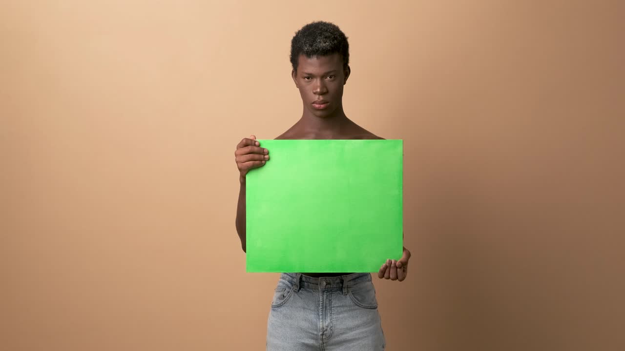Angry young black shirtless man with a green chroma banner looking at camera with beige background