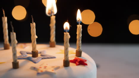 close up of four candles being lit with a match, amongst unlit candles on a white, decorated birthday cake, bokeh lights in the background