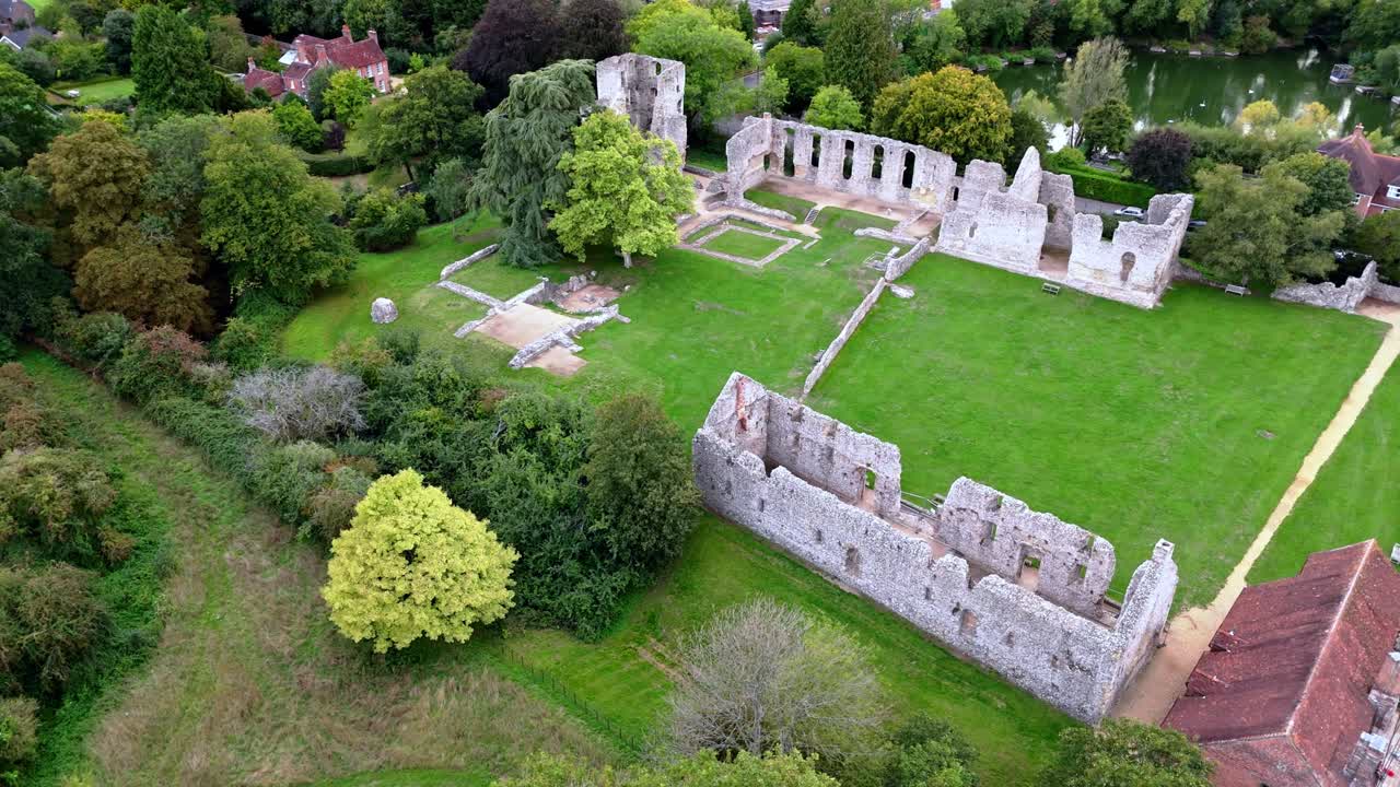 Low-angle aerial drone pans right across Bishop’s Waltham Palace ruins and pond, showcasing historic stone walls, autumn trees, and warm sunset light over the tranquil village setting