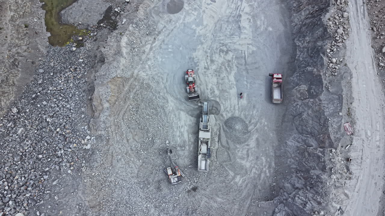 Construction vehicles work on a rocky site for a project in a quarry during daylight hours, showing heavy machinery and raw materials