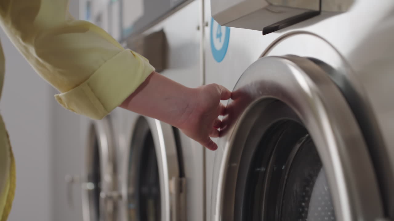 Close up laundress hand glides across labelled washer number 3, stainless drum hums while clothes tumble inside, digital controls glow, reflection shows service, hygiene in modern laundromat
