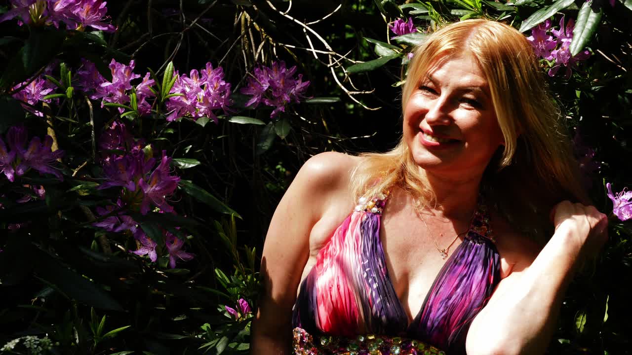 보라색 꽃을 배경으로 카메라에 미소 짓는 매력적인 여성 중간 초상화 4k 슬로우 모션