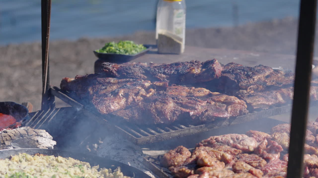 Still camera clip portraying Argentine asado type barbecue stake on fire, with ashes and smoke on the air and river in background.