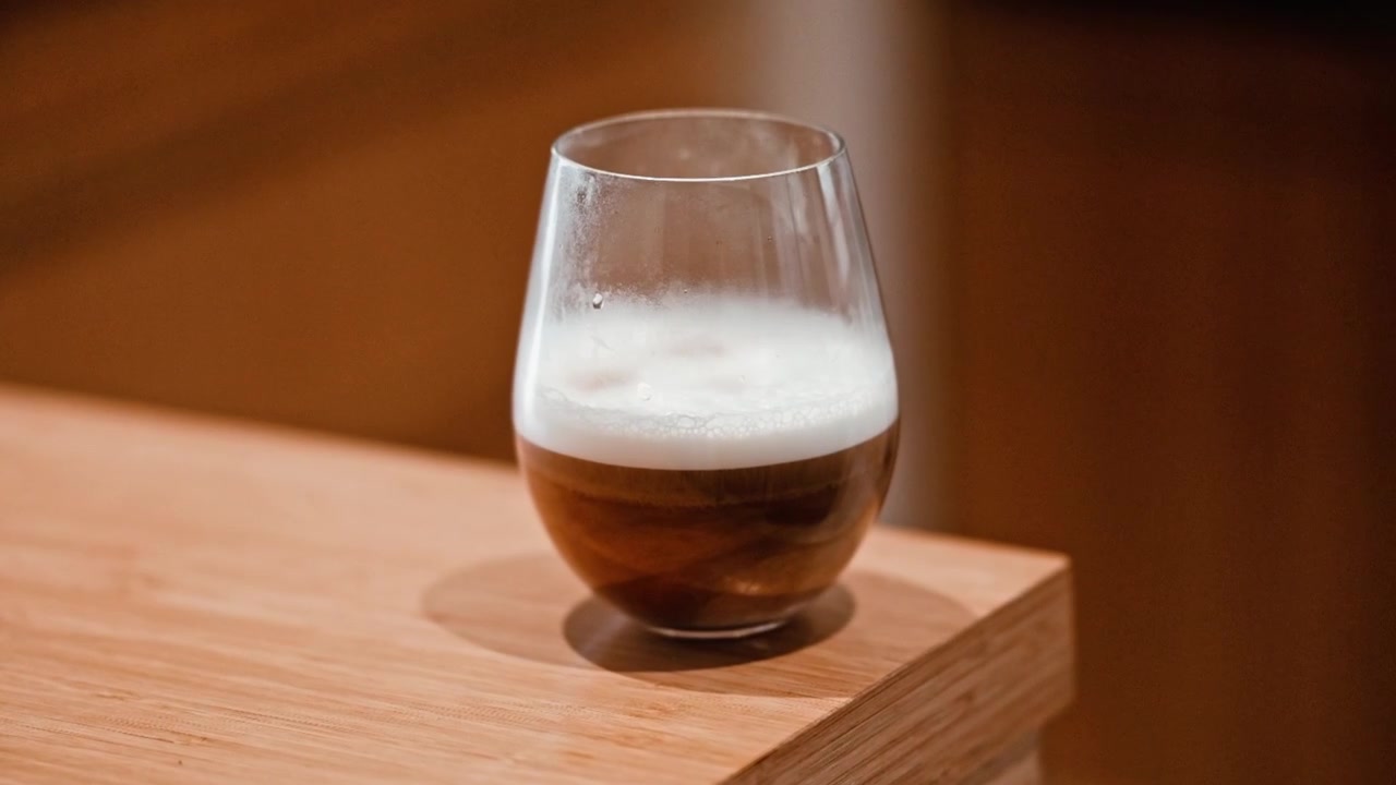 A coffee in a glass with frothed milk standing on a bamboo counter top