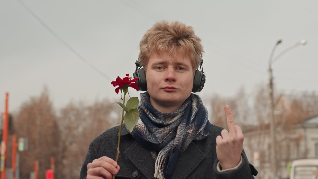 Белый мужчина в наушниках с красной розой, на фоне улицы и в шерстяном шарфе. Игривая ухмылка творца и провокационный жест. Ироничный юмор и уверенное обаяние. Откровенный городской портрет с теплым оттенком и содержанием.