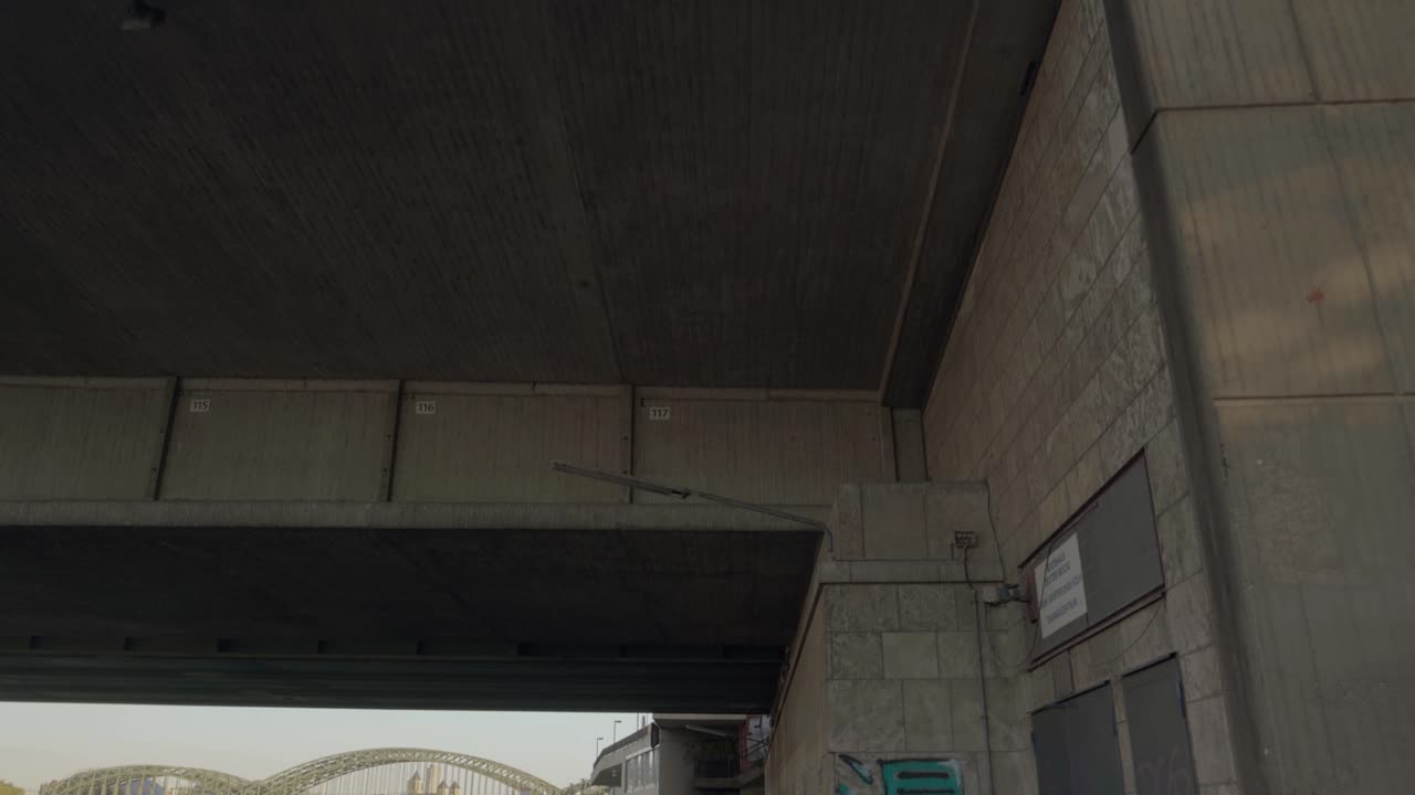 dolly hacia adelante mientras mira hacia arriba y luego se inclina hacia abajo para ver una pared pintada con graffiti y un camino sombreado debajo del puente en un día soleado a lo largo de un río