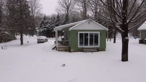 drone flying towards green cottage surrounded by snow-covered ground during snowfall in winter. sherbrooke, canada. fpv