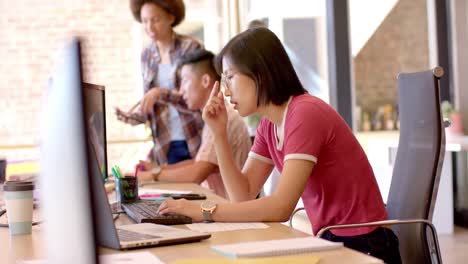 Reflexiva-Mujer-Asiática-Trabajando-En-La-Computadora,-Con-Diversos-Colegas-En-Una-Oficina-Informal,-Cámara-Lenta