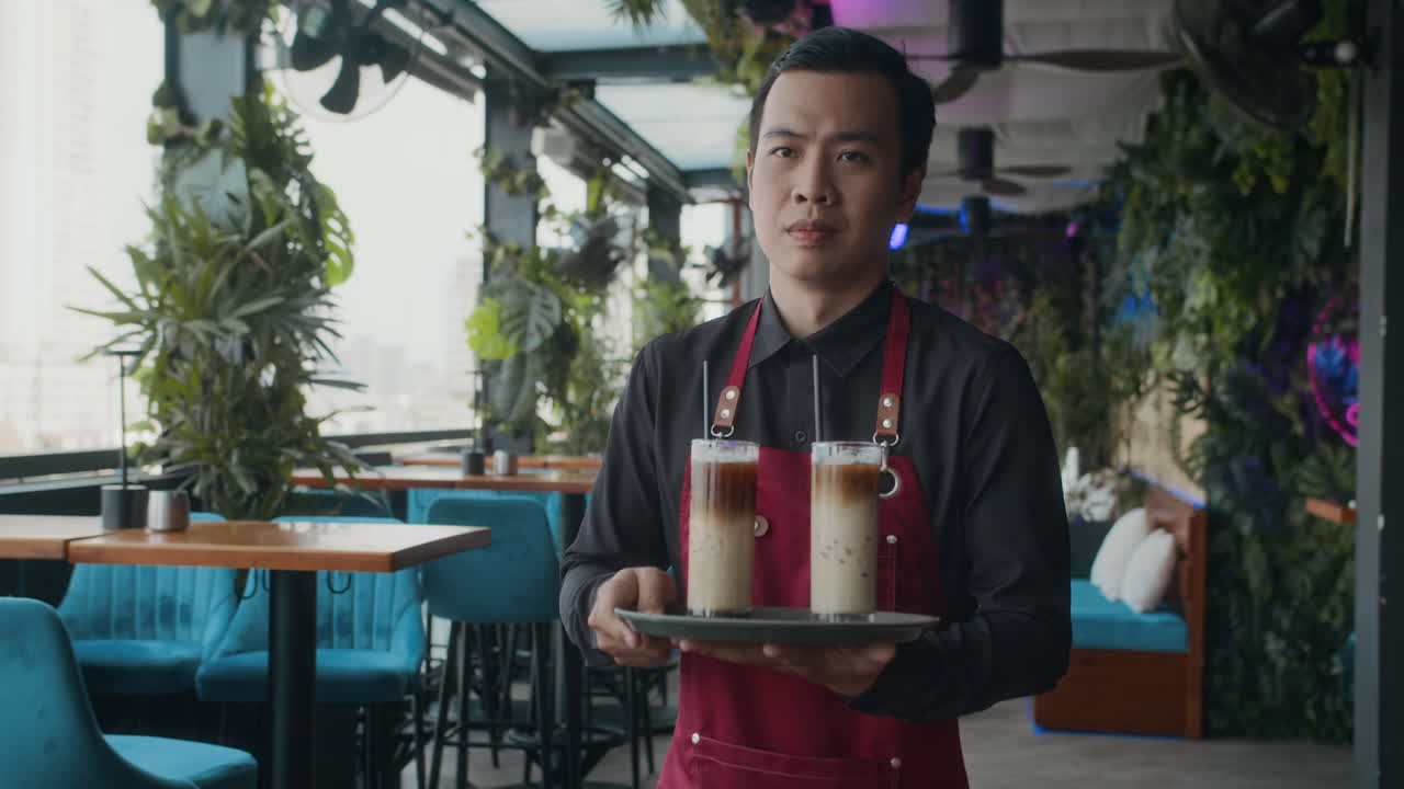 Pelayan Asia Tersenyum Membawa Baki Berisi Minuman di Kafe dengan Gerakan Lambat