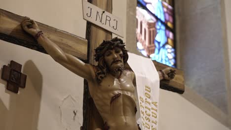 Close-up-of-a-crucifix-with-Jesus-in-a-church,-featuring-a-stained-glass-window-in-the-background