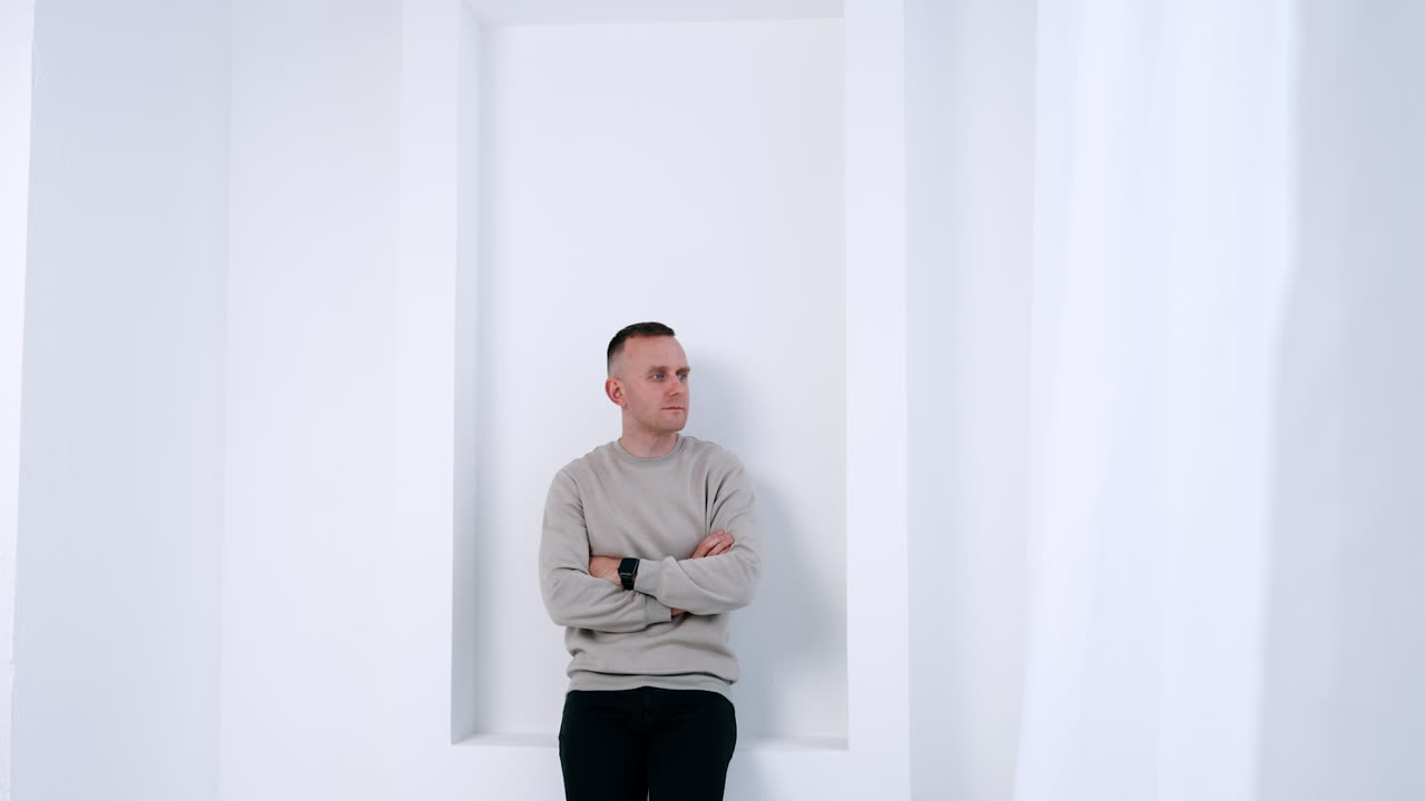 Cropped image of a man standing in the white room leaning on the wall. Man in grey sweater holding hands crossed on his chest looking aside.