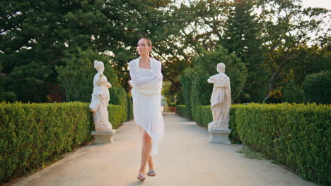 gorgeous girl going alley with ancient sculptures. smiling woman walking garden