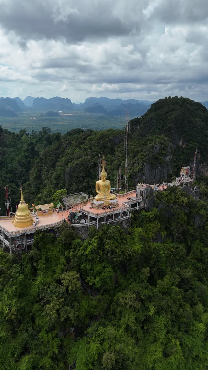 Tempio della Grotta della Tigre a Krabi, Thailandia. Statua del Buddha dorata e pagoda su una cima di montagna che domina una lussureggiante valle verde in una giornata nuvolosa, vista aerea verticale