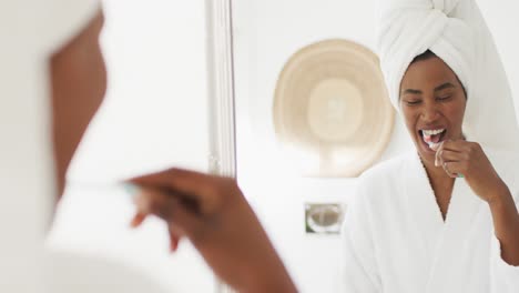 Video-Einer-Glücklichen-Afroamerikanischen-Frau-In-Robe,-Die-In-Den-Spiegel-Schaut-Und-Zähne-Putzt