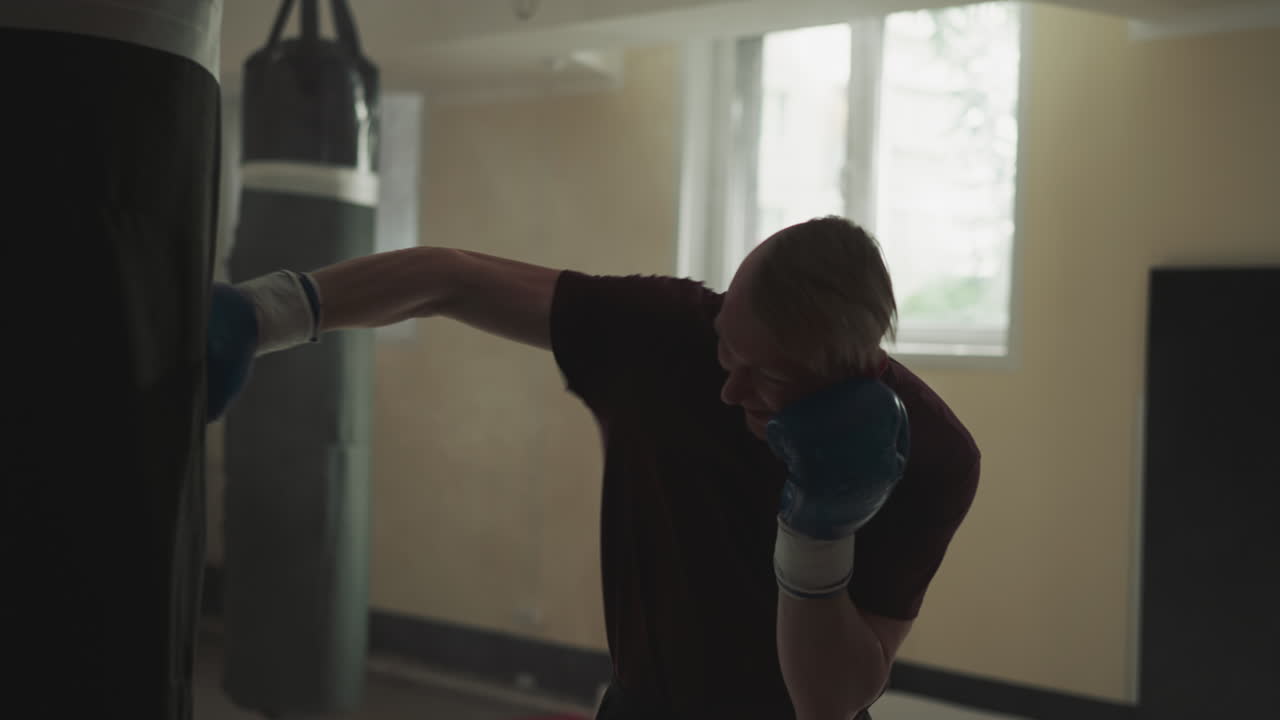 스포츠 클럽에서 싱백을 때리는 스포츠 선수, 권투 장갑을 입은 숙련된 운동선수, 체육관에서 무거운 물건을 공격하는 느린 동작, 무술 강사