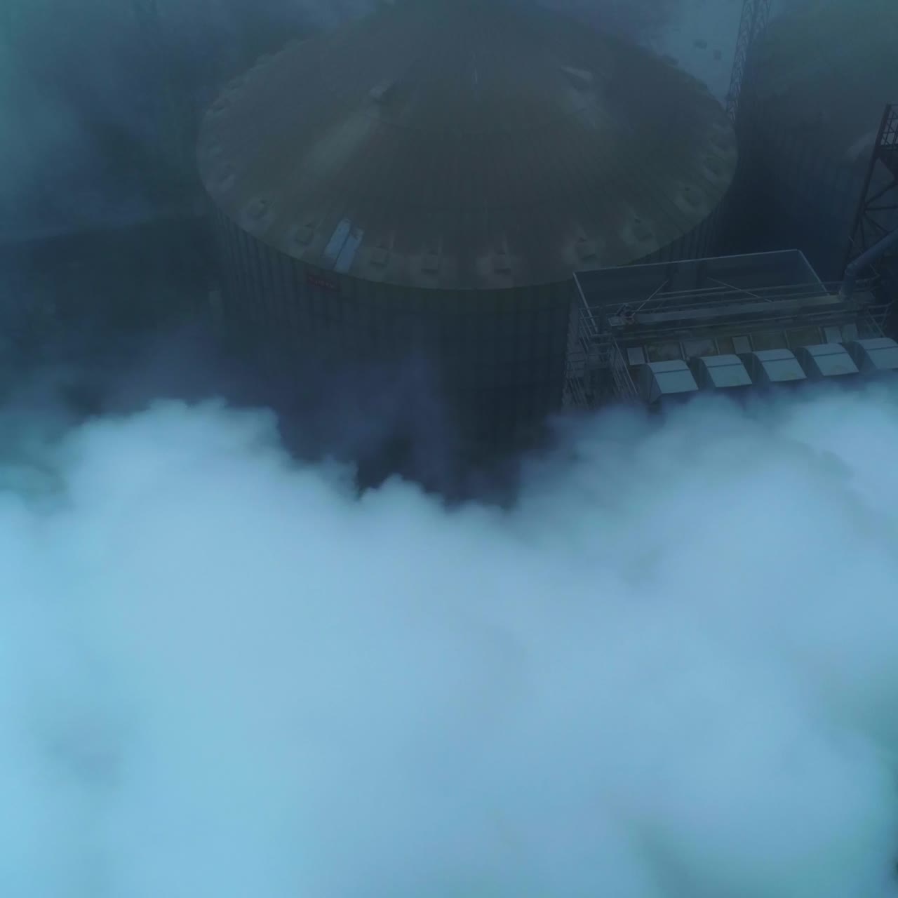 Huge functioning agribusiness plant for grain storage and processing. Top view on the silo tanks roofs in the white smoke and fog