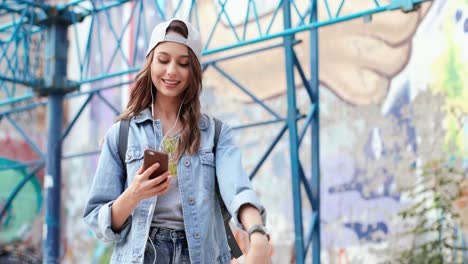 chica caucásica con gorra y auriculares escuchando música en el smartphone mientras baila y camina por la calle