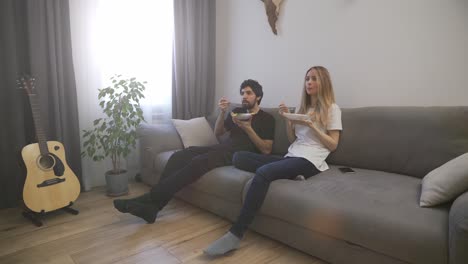 happy couple eating dinner from white bowls, sitting on the sofa