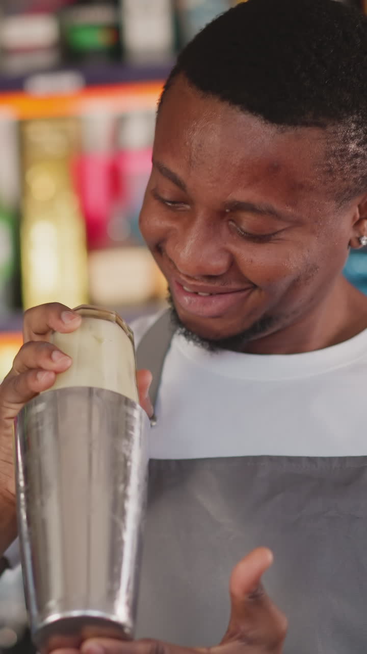 Bearded barman prepares drink with shaker in bar closeup. Joyful African American bartender shakes cocktail in metal cup with glass above in nightclub
